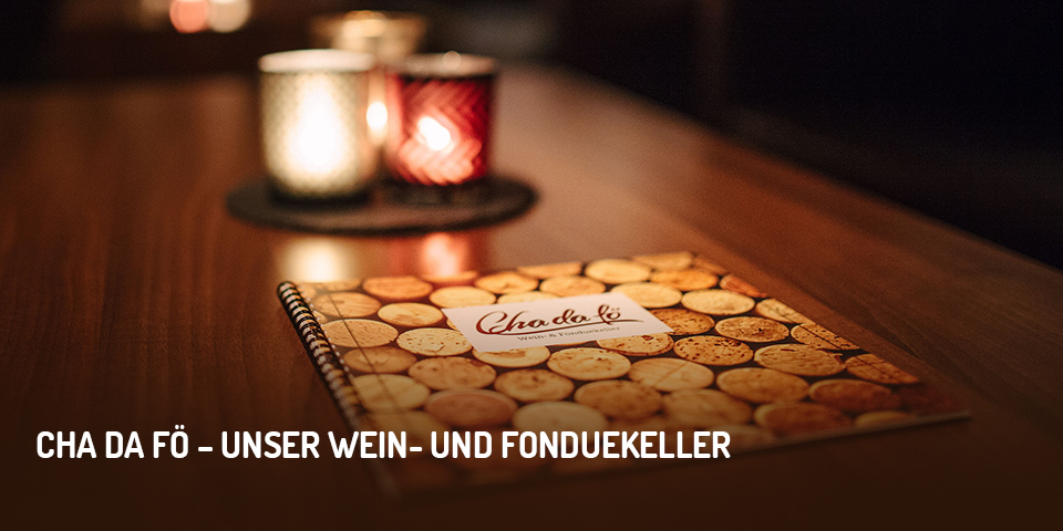 Dunkler Tisch mit drei farbigen, leuchtenden Kerzengläsern und Speisekarte des Restaurants Cha da fö, Aufschrift im Bild „Cha da fö – unser Wein- und Fonduekeller“