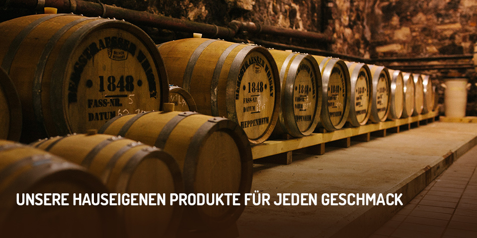 Eine Reihe von Eichenfässern, die im Gewölbekeller lagern; Bildaufschrift: „Unsere hauseigenen Produkte für jeden Geschmack“ 