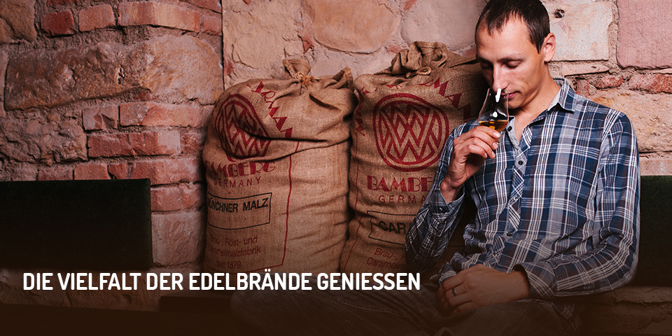 Destillateurmeister Gregor Thormann sitzt vor einer Backsteinwand neben zwei gefüllten Malzsäcken und riecht an einem Glas, das mit einer Spirituose gefüllt ist