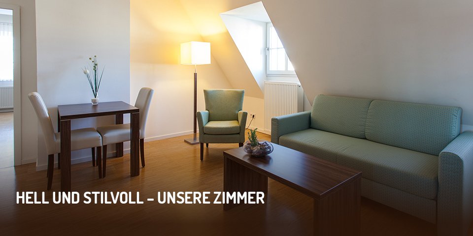 Wohnbereich eines Hotelzimmers im Halben Mond mit Dachschräge, darunter Couch, Couchtisch, leuchtende Stehlampe und Sessel, links kleiner Esstisch mit zwei Stühlen; Bild enthält Aufschrift „Hell und stilvoll – unsere Zimmer“