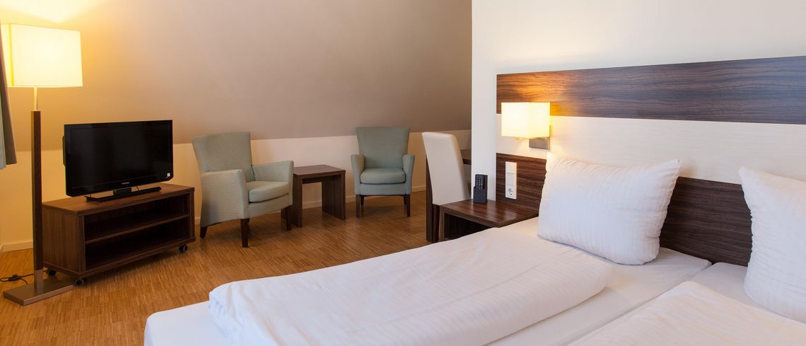 Hotelzimmer des Halben Mondes mit Doppelbett, seitlich Stehlampe, Sitzgelegenheit mit zwei Sesseln und Flachbildschirmfernseher auf kleiner Kommode