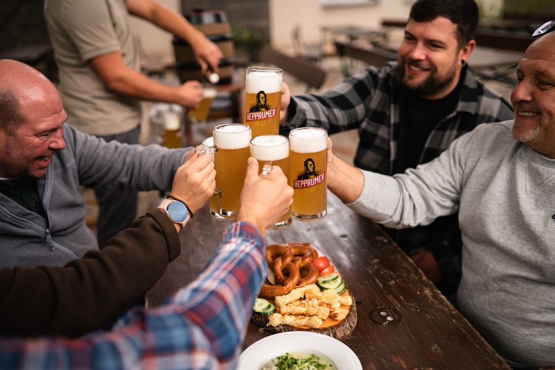 Menschen, die an einem gedeckten Biertisch mit gefüllten Hepprumer 1847-Biergläsern anstoßen, eine Person im Hintergrund zapft Bier aus einem kleinen Bierfass