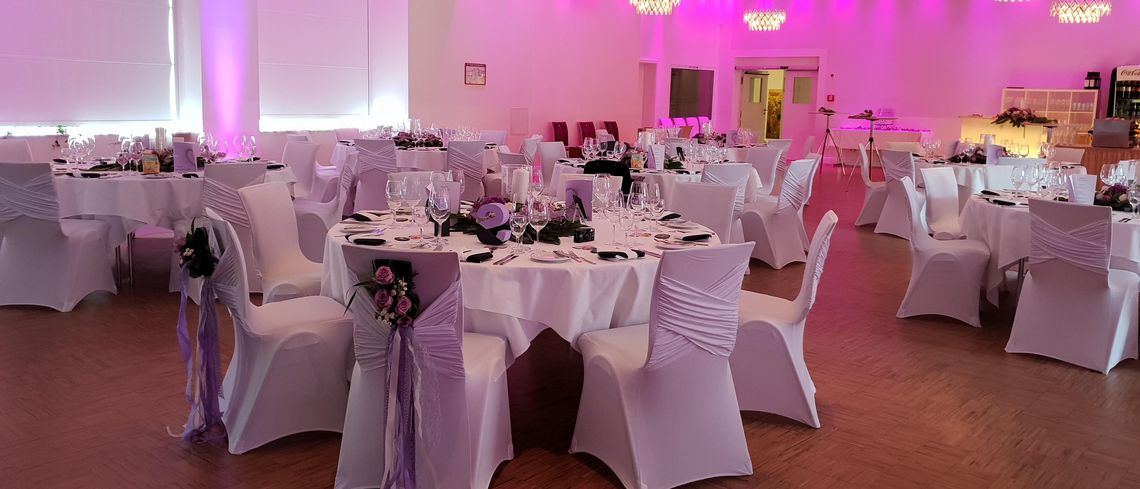 Ballsaal des Halben Mondes mit hellen, festlich eingedeckten Tischen und Stühlen sowie rosafarbene Beleuchtung der Wände, Bar im Hintergrund