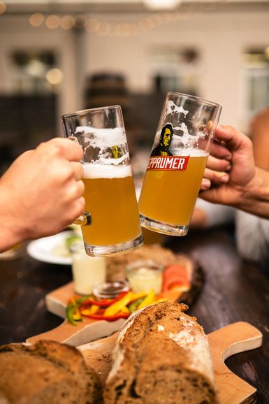 Zwei Hände über einem mit Speisen gedeckten Biertisch, die mit dem Bier Hepprumer 1847 im Glas anstoßen