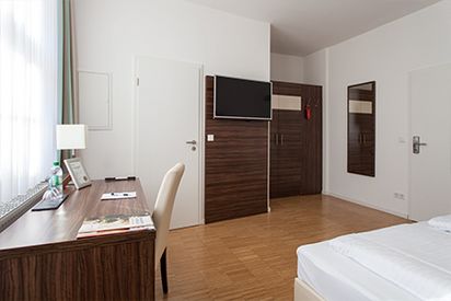 Blick in eines der Hotelzimmer des Halben Mondes, links dunkelbrauner Schreibtisch am Fenster, mittig Flachbildschirmfernseher an der Wand, rechts Teil des Bettes