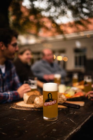 Glas Hepprumer 1847 Bier auf einem mit Speisen gedeckten Biertisch, im Hintergrund Menschen, die an diesem Tisch sitzen