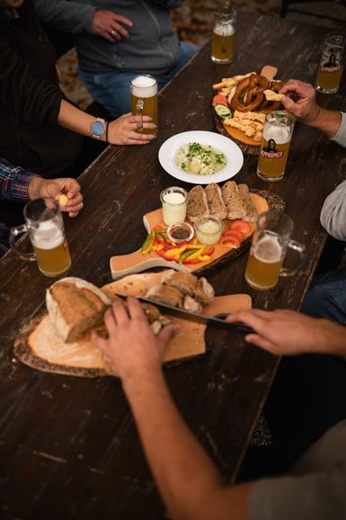 Gedeckter Biertisch, an dem mehrere Personen speisen und Bier trinken, eine davon schneidet Brot